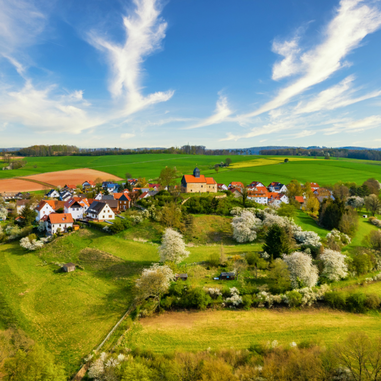 Luftaufnahme der Kapelle Schönberg an einem sonnigen Frühlingstag mit blühenden Bäumen und blauem Himmel.