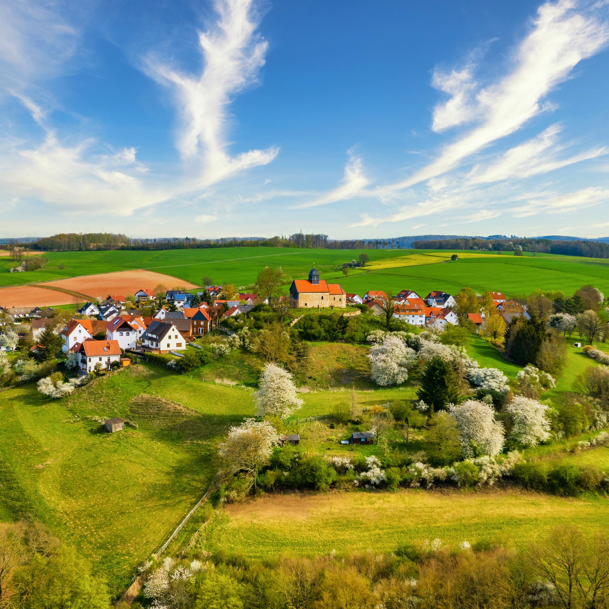 Luftaufnahme vom Schönberg mit Kapelle, umgeben von grünen Feldern, blühenden Bäumen und sanften Hügeln unter blauem Himmel mit Wolken.